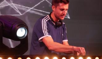 DJ Max Perth w sportowej koszulce gra za konsolą ze słuchawkami na szyi.