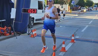 Zwycięzca biegu na 5 km Jakub Matuszczak (w niebiesko-białej koszulce i okularach przeciwsłonecznych) przekracza linię mety na drugim okrążeniu obok zegara pomiaru czasu wskazującego wynik 10:53.