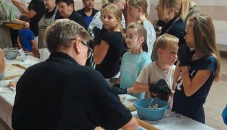 Podczas warsztatów kulinarnych kobieta w czarnej koszulce zagniata mąkę na dużej drewnianej stolnicy. Otacza ją grupa zaciekawionych dzieci i dorosłych, obserwujących proces tworzenia ciasta.