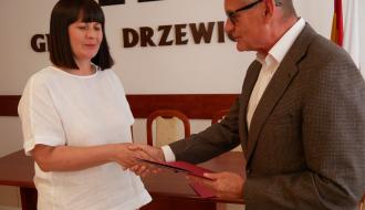  Burmistrz Drzewicy w garniturze i okularach podaje czerwoną teczkę z aktem mianowania i ściska dłoń nauczycielki w białej bluzce.