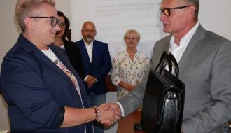 Uścisk dłoni Pani Sołtys Werówki w ciemnej marynarce i okularach oraz Burmistrza Drzewicy w siwym garniturze, trzymającego czarną torbę. W tle stoją radni oraz widać fragment ekranu projekcyjnego.