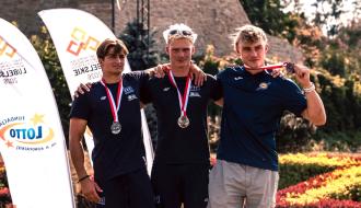 Trzech młodych kajakarzy górskich pozuje z medalami na szyi, obejmując się w pasie i za barki. Zawodnicy po lewej i w środku noszą ciemnoczarne koszulki, a zawodnik po prawej - Maciej Bartos - ciemnoniebieską koszulkę LKK Drzewica. W tle pionowe banery reklamowe.