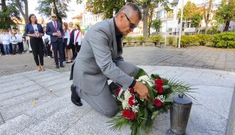 Burmistrz Drzewicy w szarym garniturze klęczy przy pomniku i składa wiązankę czerwonych i białych róż.