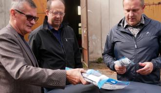 Trzech mężczyzn - Burmistrz Drzewicy i pracownicy UM w Drzewicy - rozmawia i ogląda akcesoria montażowe położone na pokrywie czarnego zbiornika na deszczówkę. 
