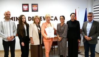 iedem osób stoi pozując do pamiątkowej fotografii w sali konferencyjnej UM w Drzewicy. Pośrodku Pani Dyrektor SP w Radzicach w jasnopomarańczowym garniturze trzyma podpisane dokumenty. W tle na białej ścianie wiszą godło Polski, herb Drzewicy oraz napis „Gmina Drzewica”. Obok stoją flagi.