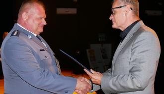 Burmistrz Drzewicy w szarym garniturze i okularach uśmiecha się, podając dłoń asp. szt. Pawłowi Janikowi w garnizonowym mundurze. Burmistrz wręcza dokument w niebieskiej okładce. Wydarzenie odbywa się na scenie.