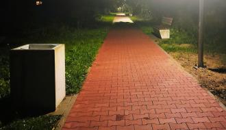 Chodnik z czerwonej kostki brukowej nocą. Po lewej stronie stoi szary, betonowy kosz na śmieci. Dalsza część ścieżki ginie w mroku i lekkiej mgle, w oddali kolejne punkty świetlne oraz ławka parkowa po prawej stronie.