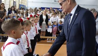 Burmistrz Drzewicy w ciemnym garniturze i okularach uśmiecha się podczas pasowania na ucznia, podając rękę chłopcu w galowym stroju z czerwonymi szelkami i muszką. W tle widać rządek uczniów oraz uśmiechniętą widownię. 