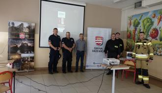 Grupa sześciu mężczyzn (policjanci, strażacy, cywil) stoi w rzędzie w sali prelekcyjnej. W środku baner Gminy Drzewica. 