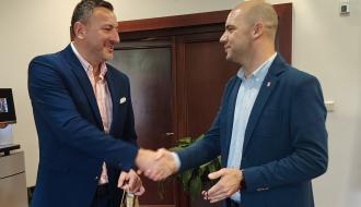 Burmistrz Nałęczowa i Wiceburmistrz Drzewicy w garniturach podają sobie dłonie i uśmiechają się, trzymając torby prezentowe w jasnym wnętrzu. 