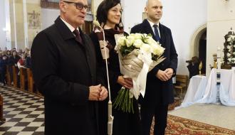 Stojąca przed ołtarzem delegacja Gminy Drzewica. Po lewej stronie Burmistrz Drzewicy w ciemnym płaszczu i okularach przemawia do mikrofonu, w środku Przewodnicząca Rady Miejskiej w Drzewicy trzyma duży bukiet białych róż, a po prawej stronie stoi Wiceburmistrz Drzewicy w garniturze.
