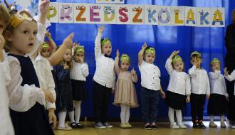 Dzieci stoją w rzędzie z uniesionymi rękami pod dekoracją z napisem „PASOWANIE NA PRZEDSZKOLAKA”.