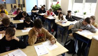 Uczniowie ze starszej kategorii wiekowej siedząc w ławkach w sali lekcyjnej biorą udział w pisemnej części turnieju