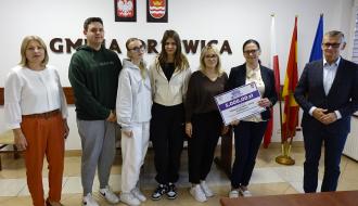 Na tle ściany z napisem GMINA DRZEWICA, godłem Polski i herbem Drzewicy od lewej stoją: Dyrektor ZEAS Małgorzata Kuchenbecker, troje uczniów i dwie nauczycielki ZSP w Drzewicy (jedna trzyma plastikową promesę), Burmistrz Drzewicy Janusz Reszelewski.