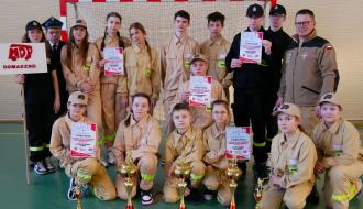  Młodzieżowe Drużyny Pożarnicze z OSP Domaszno wraz z opiekunami prezentują zdobyte trofea. Dziesięć osób stoi, siedem klęczy. W tle bramka do piłki ręcznej.