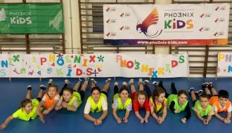W hali sportowej dziesięcioro dzieci leży na parkiecie przodem do fotografa. W tle drabinki i banery Pho3nix Kids
