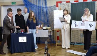 Turniej wiedzowy w hali sportowej SP w Brzustowcu. Po lewej stronie „Drużyna B” (troje nastolatków i Burmistrz Drzewicy) stoi przy stole z czerwonym przyciskiem. Po prawej prowadząca czyta z notatek, a druga nauczycielka prezentuje planszę z grafikami terminala, dokumentu i paczki. W tle widać wyposażenie sportowe i tablice z punktacją.