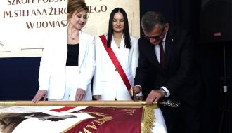 Trzy osoby w strojach formalnych skupione przy sztandarze podczas uroczystości nadania sztandaru SP im. Stefana Żeromskiego w Domasznie.