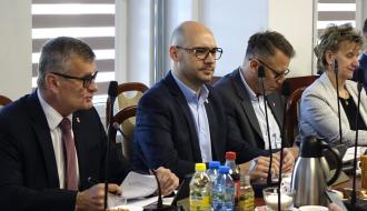 Przy stole z mikrofonami i dokumentami siedzą cztery osoby w formalnych strojach – Burmistrz i Zastępca Burmistrza Drzewicy oraz dwoje radnych. Głos zabiera Burmistrz Drzewicy, pozostali słuchają. Na stole widoczne są napoje i materiały do pracy.