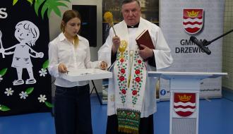 Ks. Wiesław Mazur w białej komży ze stułą trzyma w dłoni kropidło oraz czerwoną księgę. Obok niego stoi nastolatka w białej koszuli, trzymająca białą tacę z naczyniem na wodę święconą. W tle baner z herbem Gminy Drzewica.