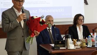Burmistrz Drzewicy w szarym garniturze i okularach przemawia do mikrofonu, trzymając bukiet czerwonych i białych róż. W tle siedzą członkowie prezydium Rady, a na ekranie wyświetlana jest treść uchwały i nazwisko Janusz Reszelewski.