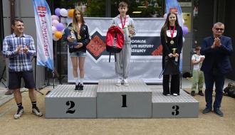  Dekoracja młodzieży na podium przed RCK w Drzewicy. Na najwyższym stopniu chłopiec w szarym dresie z pucharem i czerwonym plecakiem. Na drugim i trzecim miejscu dziewczęta z pucharami i nagrodami. Po bokach asystują Wiceprezes LGD „Nad Drzewiczką” i Burmistrz Drzewicy, klaszcząc w dłonie. W tle baner RCK i flagi promocyjne.