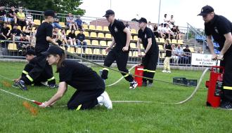 Start młodzieżowej drużyny pożarniczej w zawodach na drzewickim stadionie. Na pierwszym planie druhna w czarnym stroju klęczy na trawie, celując prądownicą węża strażackiego. Obok niej młodzi druhowie obsługują ręczne, czerwone hydronetki, pompując wodę. W tle żółte trybuny stadionu z siedzącymi widzami.