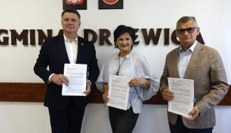 Członek Zarządu Woj. Łódzkiego Artur Bagieński, Teresa Szklarczyk i Burmistrz Drzewicy Janusz Reszelewski stoją w rzędzie na tle ściany z napisem „GMINA DRZEWICA”, godłem i herbem. Każda z osób trzyma w dłoniach i prezentuje podpisaną stronę dokumentu, patrząc w stronę obiektywu.