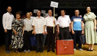Grupa dziewięciu osób pozuje do zdjęcia na scenie w sali widowiskowej RCK. Mężczyzna w centrum trzyma tabliczkę z napisem „WTZ Drzewica”. Uczestnicy są ubrani w stroje galowe oraz codzienne. Na pierwszym planie, przed grupą, stoi duża, czerwona torba prezentowa z logo „Gerlach”. W tle inne tabliczki informacyjne.