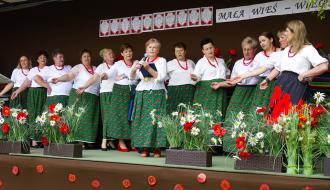 Występ artystyczny na scenie plenerowej. Grupa kilkunastu kobiet w strojach ludowych (białe bluzki, zielone spódnice w maki i czerwone korale) śpiewa, wykonując gesty dłońmi. Jedna z nich trzyma mikrofon. Nad nimi wisi napis „ŚWIERCZYNA MAŁA WIEŚ”. Przód sceny udekorowany jest sztucznymi makami i rumiankami.