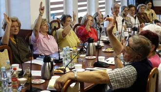 Grupa seniorów siedzących przy stole konferencyjnym bierze udział w głosowaniu, podnosząc ręce. Na stole stoją napoje, szklanki oraz talerze z ciastkami.
