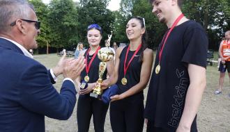 Burmistrz Drzewicy w garniturze klaszcze, gratulując trójce sportowców - dwóm kobietom i mężczyźnie - trzymających złoty puchar za drugie miejsce. W tle drzewa.