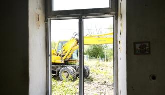 Widok z wnętrza starego, pustego pomieszczenia w „Ceglance” przez duże okno na stojącą na zewnątrz żółtą koparkę Komatsu
