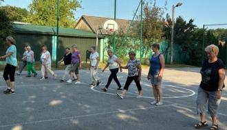 Seniorki uczestniczące w zajęciach ruchowych, maszerujące w rzędzie po otwartym boisku sportowym