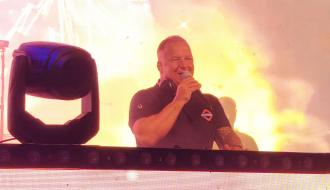 DJ Hubertuse stoi z mikrofonem na scenie z jasnym podświetleniem w tle.