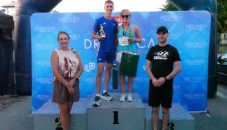 Najlepsi biegacze Gminy Drzewica w kategorii kobiet i mężczyzn – Natalia Pacierpnik i Jakub Matuszczak - stoją na podium na 1. miejscu z pucharami, w otoczeniu organizatorów na tle ścianki „Drzewica Poczuj Pasję”.