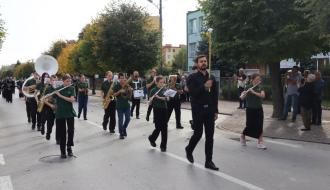 Przemarsz orkiestry dętej Gminy Rozprza w zielonych koszulkach polo. Na czele idzie dyrygent w czarnej koszuli z buławą, za nim muzycy grający na fletach poprzecznych, saksofonach i sousafonie. 
