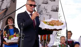 Burmistrz Drzewicy w garniturze i okularach przeciwsłonecznych przemawiający do mikrofonu na scenie, trzymając ozdobny chleb dożynkowy.