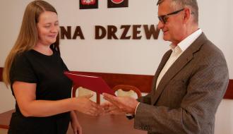 Burmistrz Drzewicy w garniturze i okularach podaje czerwoną teczkę z aktem mianowania i ściska dłoń nauczycielki w czarnej sukience.