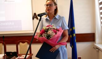 Pani Dyrektor Przedszkola Samorządowego w Drzewicy w błękitnej sukience i okularach stoi przy mikrofonie, trzymając w rękach bukiet różowych kwiatów oraz granatową teczkę. W tle stoi flaga Unii Europejskiej.