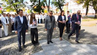  Delegacja władz samorządowych Gminy Drzewica i młodzieży stoi w skupieniu przed Pomnikiem Pamięci po złożeniu kwiatów i zniczy. Po prawej stronie w tle widać sztandar oraz uczniów. Słońce oświetla płytę pomnika i uczestników uroczystości.
