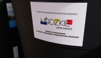Zbliżenie na białą tabliczkę informacyjną naklejoną na czarnym pojemniku. Na tabliczce widnieje napis: „Urząd Marszałkowski Województwa Łódzkiego. Łódzkie Łapie Deszcz. Zadanie finansowane z budżetu Samorządu Województwa Łódzkiego”.