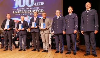 Wyróżnieni w garniturach oraz wyżsi rangą funkcjonariusze Policji stoją w jednym rzędzie na drewnianej scenie. Czterej z nich trzymają pamiątkowe statuetki. W tle widnieje duży ekran z napisem „100-lecie powołania funkcji dzielnicowego w Polskiej Policji” oraz godłem.