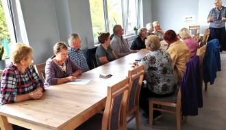 Kobieta w paski prowadzi warsztaty dla grupy seniorów siedzących przy długim stole w nowoczesnej sali świetlicy w Idzikowicach. 