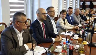 Przy stole siedzą uczestnicy sesji. Pierwszy z lewej Burmistrz Drzewicy w okularach i szarym garniturze przemawia do mikrofonu. Obok niego siedzą Wiceburmistrz, radni a dalej dyrektorki szkół.