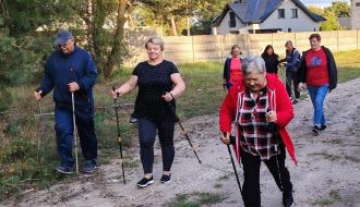 Grupa seniorów spaceruje z kijami do nordic walking po gruntowej drodze w pobliżu zabudowań.