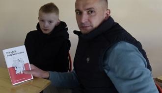 Mężczyzna i chłopiec siedzą przy stole i patrzą w stronę aparatu. Mężczyzna trzyma czerwoną broszurę „Poradnik bezpieczeństwa”.