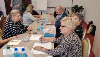 Uczestnicy spotkania siedzą przy długich stołach podczas warsztatów. Na pierwszym planie kobieta w bluzce w panterkę robi notatki. 