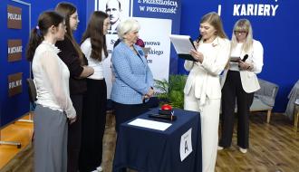 Turniej wiedzowy w hali sportowej SP w Brzustowcu. Po lewej stronie trzy uczennice i Pani Wojewoda Łódzki w niebieskim żakiecie stoją przy stole „Drużyny A” z czerwonym przyciskiem. Po prawej prowadząca w białym garniturze czyta tekst do mikrofonu, a obok niej stoi druga nauczycielka. W tle widoczna jest niebieska ścianka z napisami.