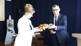 Kobieta w białym garniturze i mężczyzna w ciemnym garniturze wymieniają się książką przewiązaną złotą wstążką podczas uroczystości w SP im. Stefana Żeromskiego w Domasznie.